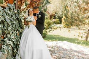 A stylish groom in a black suit and a cute bride in a white dress with a long veil are hugging in a park. Wedding portrait of smiling and happy newlyweds. photo