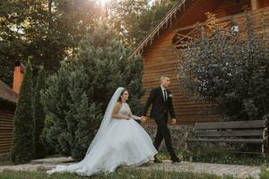 A stylish groom in a black suit and a cute bride in a white dress with a long veil are hugging and walking near green tall trees. Wedding portrait of smiling and happy newlyweds. photo