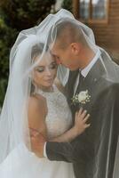 gorgeous happy luxurious brunette bride and elegant stylish groom stand under a veil and embrace tenderly photo