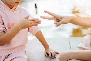 dos niña amigos son jugando un juego de papel tijeras. caucásico niños sentado por el fuente al aire libre, jugando juntos. un interesante y entretenido actividad para niños. foto