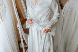 Happy dark-haired brunette bride in a satin robe and professional make-up standing in her room near the mirror and getting ready for her wedding day photo