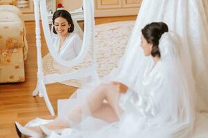 contento morena novia en blanco satín vestido con largo velo profesional maquillaje y cabello. el novia es sentado en el habitación en el piso cerca el espejo. Boda vestir en un maniquí foto