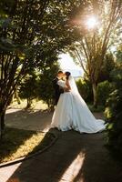 Warm sunny portrait of a happy bride with a bouquet of orchids in her hands and a groom in a classic suit, at sunset. Warm summer time. A long veil, a luxurious white dress with a train. photo