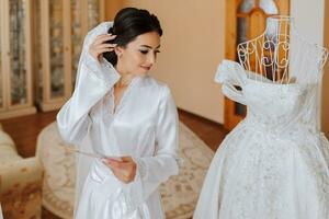 A beautiful bride holds a letter from the groom. The bride in a white satin robe is standing next to the dress on a mannequin, reading a letter from her lover. Wedding vows. Morning of the bride. photo