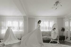 Happy dark-haired brunette bride in a satin robe and professional make-up standing in her room near the mirror and getting ready for her wedding day photo