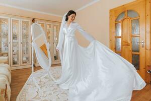 Happy dark-haired brunette bride in a satin robe and professional make-up standing in her room near the mirror and getting ready for her wedding day photo