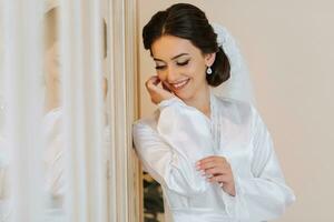 happy brunette bride in white robe with long veil professional makeup and hairstyle in bedroom. photo