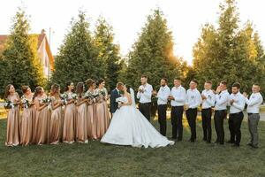 a gorgeous bride with a bouquet of orchids and a stylish groom pose in a sunny garden with bridesmaids and groomsmen on their wedding day. luxury wedding couple having fun with friends photo