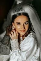 hermosa novia con largo oscuro Rizado pelo en un largo blanco túnica en el Mañana. el novia es preparando para el boda. profesional maquillaje y cabello. un corona en el cabeza y un largo velo de el novia foto