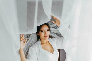 retrato de un niña en un Mañana túnica. contento novia con profesional maquillaje y largo velo posando para fotógrafo. el novia es preparando para el Boda foto