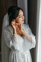 Portrait of a girl in a dressing gown. Happy bride with professional make-up and long veil poses for photographer. The bride puts on earrings and prepares for the wedding photo