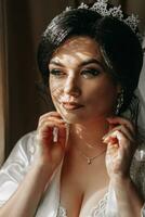 Portrait of a bride woman in a white robe with a beautiful hairstyle near the window, looking to the side. Sensual cute bride posing. The concept of luxury among fashionable style. photo