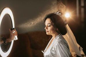 un hermosa joven mujer con oscuro cabello, un Boda peinado, un corona en su cabeza es haciendo maquillaje para un Boda o un foto disparo. el trabajo de un maquillaje artista. Mañana de el novia. profesional maquillaje