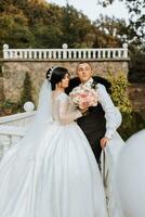 a beautiful bride in a chic wedding dress and an elegant groom stand on ancient steps in the park photo