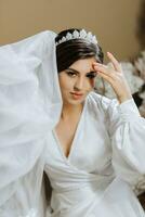 Portrait of a beautiful brunette woman in a glamorous silk wedding gown, a crown on her head, and a long veil. Posing for a photographer. photo