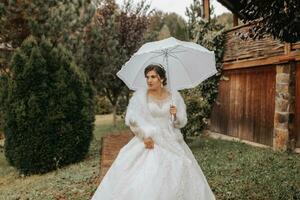 Beautiful stylish bride with a white umbrella, walks in the rain in the park, wedding photo session in the rain. A model with an umbrella in a lush wedding dress