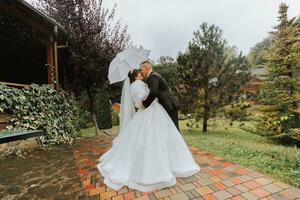 novia y novio con un blanco paraguas, camina en el lluvia en el parque, Boda foto sesión en el lluvia. un modelo con un paraguas en un magnífico Boda vestido. el novio Guías el novia por el mano