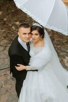 novia y novio con un blanco paraguas, camina en el lluvia en el parque, Boda foto sesión en el lluvia. un modelo con un paraguas en un magnífico Boda vestido. el novio Guías el novia por el mano