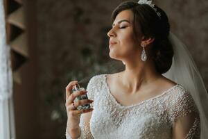 Female beauty. Cute woman at home. The bride sprays perfume on her body. A stylish woman wears a white dress. Spray delicate perfume. Stylish glass bottle of perfume in hands. photo