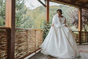 A beautiful queen in a wedding dress. charming woman bride walking in the park. The wedding outfit is a white lush dress, a luxurious raincoat, a white cape made of swan feathers. photo