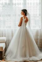 The bride in a chic lush white dress with bows on her shoulders is standing by the window in a beautiful room. photo