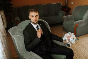 portrait. A stylish man with a beard in a white shirt, a classic suit is sitting, posing on a sofa, holding a bouquet. A stylish watch. Men's style. Fashion. Business photo