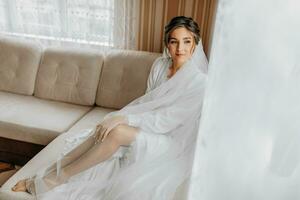 A stunning young bride poses by the window holding her veil in her hands. A beautiful woman of Caucasian appearance in an elegant white robe and luxurious hairstyle. Wedding makeup photo