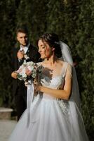 beautiful elegant luxury bride with veil and stylish groom walking on street near tall green trees photo