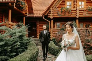 beautiful elegant luxury bride with veil and stylish groom walking on the street near tall autumn trees and wooden houses photo