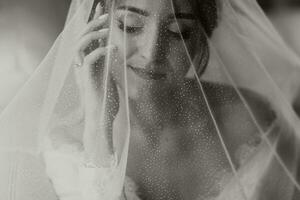 un maravilloso joven novia poses por el ventana participación su velo en su manos. un hermosa mujer de caucásico apariencia en un elegante blanco túnica y lujoso peinado. Boda maquillaje foto