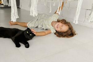 un contento niño es acostado en el veranda con su favorito negro gato. un pequeño niña descansa en el piso de el veranda en un soleado verano día. un contento y despreocupado infancia. foto