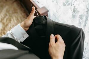 the man holds his black jacket on a hanger in his hands. The groom is preparing for the wedding ceremony. Detailed close-up photo of hands