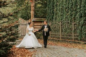 A happy couple of stylish newlyweds in a green park on an autumn day. The bride in a long white dress and the groom in a black suit walk forward. Wedding day. photo