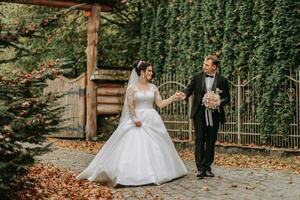 A happy couple of stylish newlyweds in a green park on an autumn day. The bride in a long white dress and the groom in a black suit walk forward. Wedding day. photo