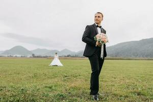 A handsome groom in a black suit and a black bow tie stands outdoors in a wide field against a background of mountains. Wedding portrait. A man in a classic suit in the foreground. photo