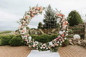 moderno ceremonia en europeo estilo. Boda arco con rosas en el antecedentes de un decorativo jardín. joyería desde Fresco flores, flores y cristales frente vista. foto