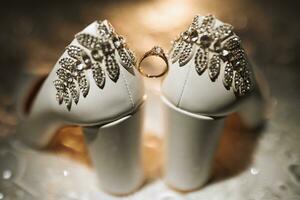 White elegant shoes. A diamond engagement ring sits between a pair of classic wedding shoes. Fashion. Style. Wedding photo