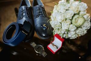 Accessories for the groom's wedding day. Blue leather shoes, belt, cufflinks, wristwatch, gold rings, bouquet of white roses on glass table. Men's fashion photo