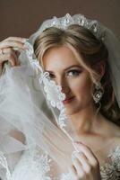 A beautiful, smiling blonde bride in a white dress is standing in her room and looking at the camera. Wedding portrait of a cheerful girl. photo