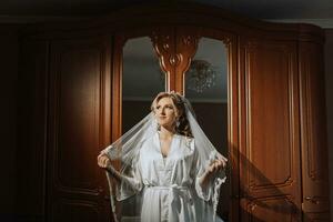 Preparation for the wedding. Beautiful young bride in a white robe in the room. A luxurious model near a mirror with a veil and a tiara on her head. Morning of the bride photo