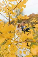 Groom and bride in autumn forest, wedding ceremony, side view. Groom and bride on the background of yellowed autumn leaves. The photo was taken through the yellowing leaves of the trees
