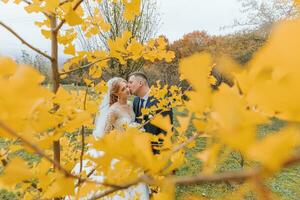 Groom and bride in autumn forest, wedding ceremony, side view. Groom and bride on the background of yellowed autumn leaves. The photo was taken through the yellowing leaves of the trees