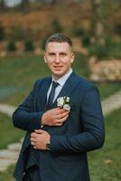 A handsome groom in a blue suit and tie is standing outdoors. Wedding portrait. A man in a classic suit photo