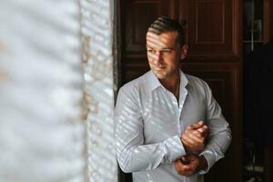 Handsome man putting on shirt standing near window at his room in morning. Preparing for some event or new workday. New opportunities, dating, wedding day or getting ready for job interview concept. photo