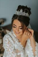 Beautiful young bride wearing earrings before wedding ceremony at home. The bride in a white robe puts on earrings in her room in the morning photo