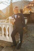 A handsome groom in a blue suit and black tie is standing outdoors. Wedding portrait. A man in a classic suit against the background of autumn leaves photo