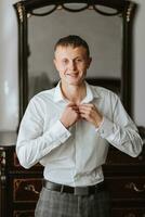 Handsome man putting on shirt standing near window at his room in morning. Preparing for some event or new workday. New opportunities, dating, wedding day or getting ready for job interview concept. photo