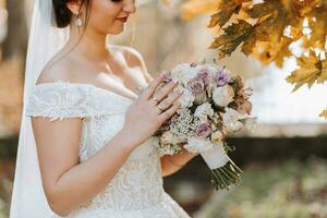 A beautiful bride stands in nature near the autumn leaves of trees with a bouquet of flowers. Close-up wedding portrait of a young bride. Wedding photography. photo