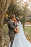 A fabulous romantic newlywed couple is hugging at sunset in an autumn park with leaves from trees on the grass photo