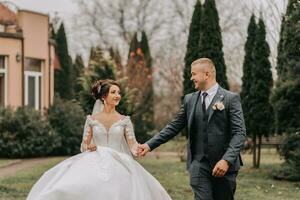 Groom and bride walking and holding hands in the park among tall green trees. Wide-angle autumn photo. Voluminous wedding dress. Classic suit of the groom photo